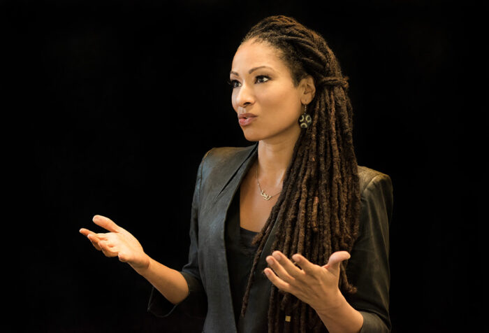 woman in long braids, black shirt and jacked gestures with her hands
