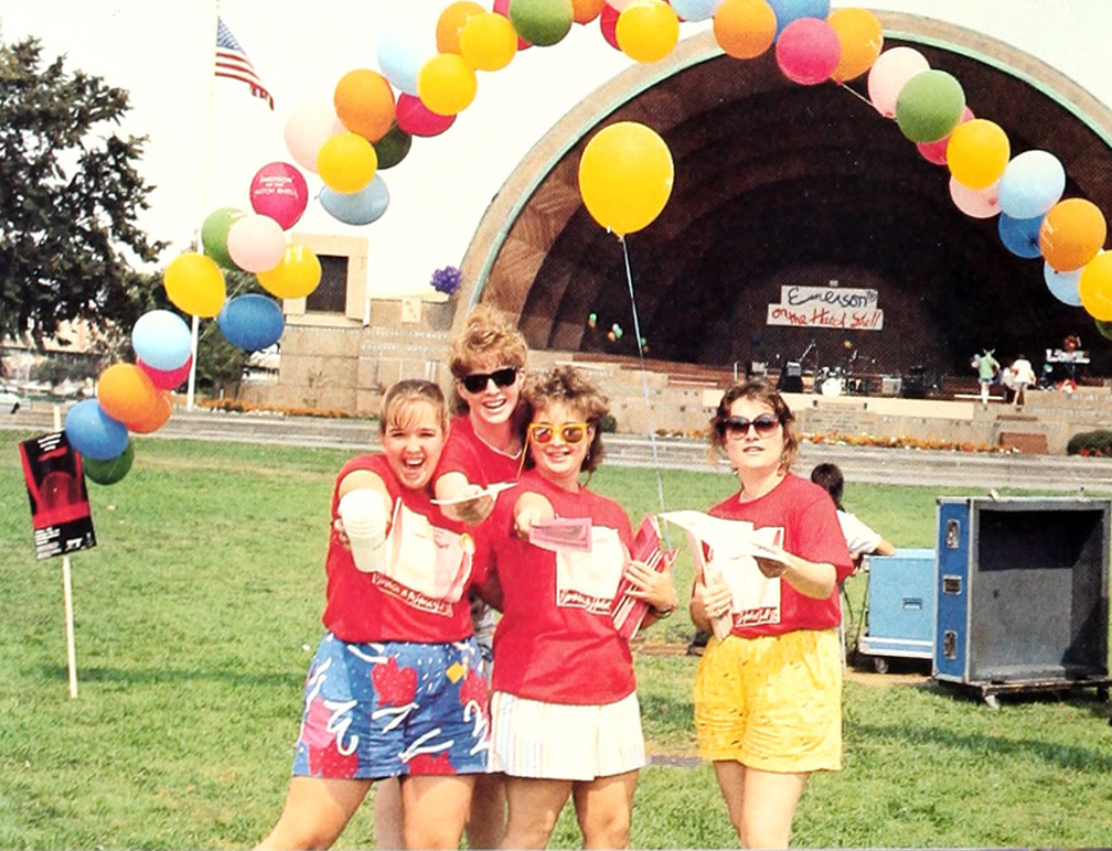 Four students in front of the Hatch Shell with balloons above them, hand out flyers.