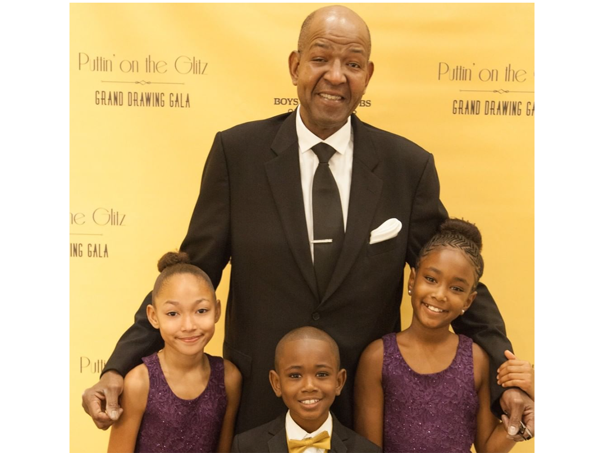 A tall man stands in a suit with his arms around three children in front of him