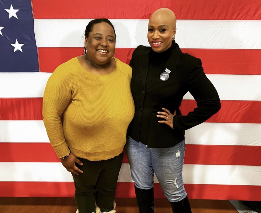 Two women stand together in front of an American flag background
