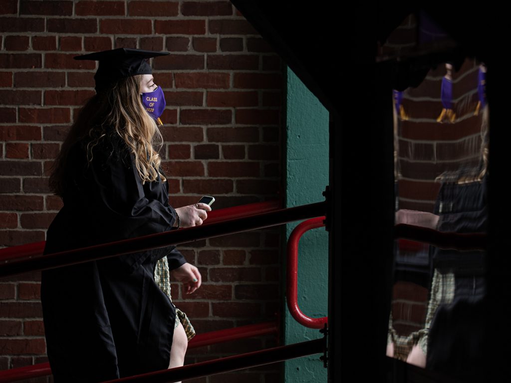 woman in cap and gown and mask walks up ramp in Fenway with phone in hand