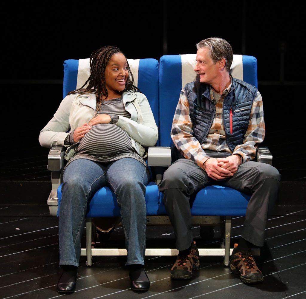 Two people act on a stage sitting in airplane seats. The woman is pregnant and the man is not. They are smiling at each other.