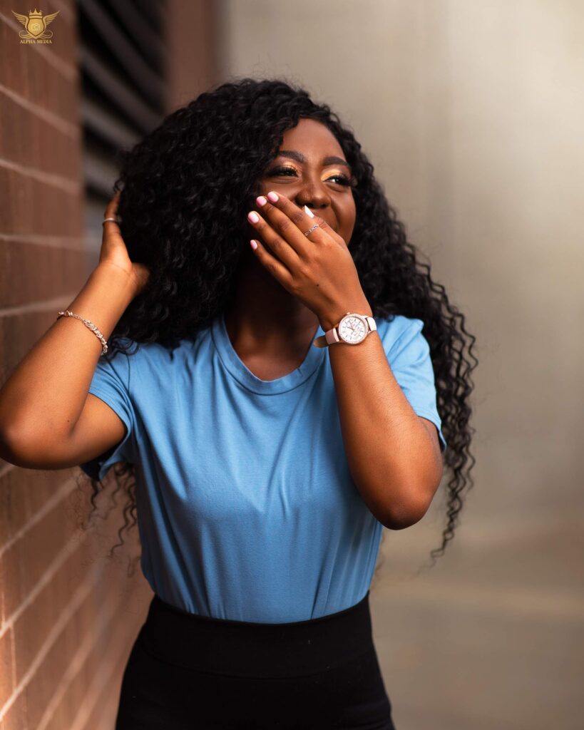 Sharon Boateng, in blue t-shirt, covers her mouth as she laughs in front of brick wall. 