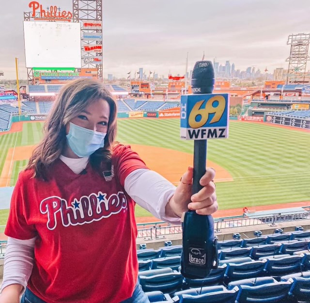 Reid wears a Phillies shirt and holds a microphone that says 69 WFMZ. She stands in a stadium in front of a baseball field. It is Citizens Bank Park.
