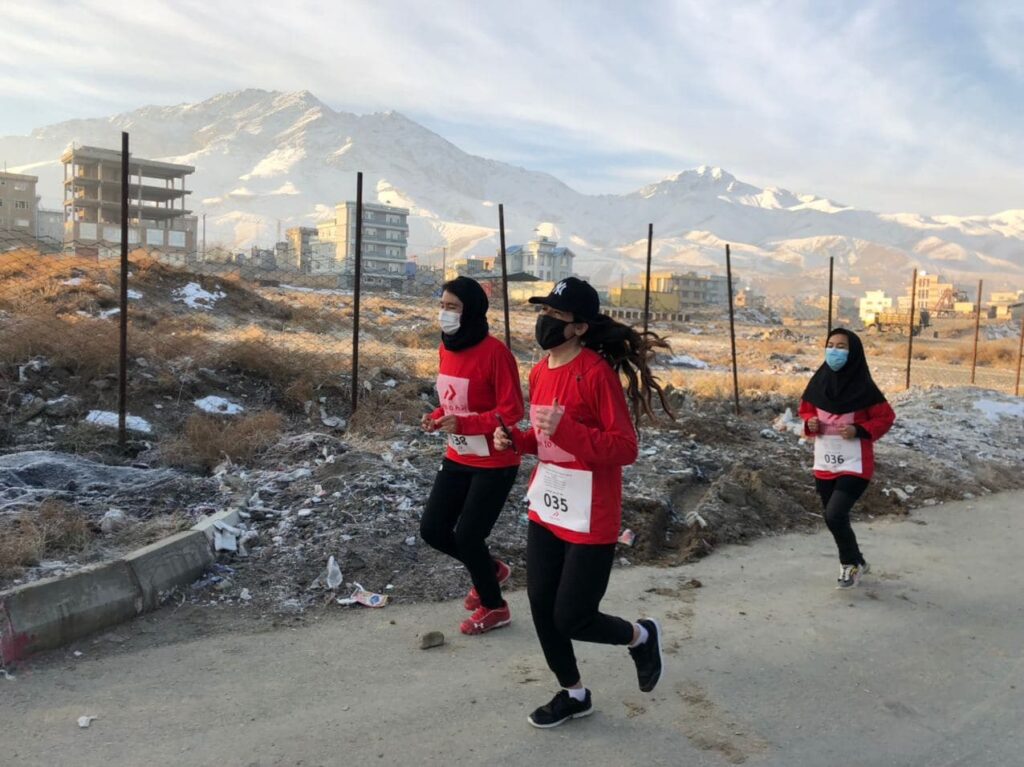 Three women run with snow capped mountains and buildings in the background