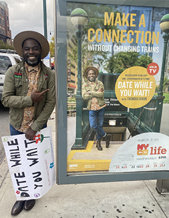 Thomas Knox stands by and points to an ad for his new show at an NYC bus stop. He holds a sign reading "Date While You Wait."
