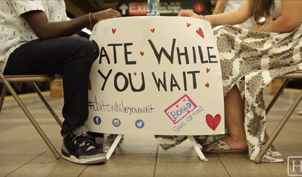 Knox and his guest are shown sitting across from each other at a small table, with a white banner reading "Date While You Wait." Their faces aren't shown, only the lower halves of their bodies, and emphasis is put on the sign between them. The sign also features the #datewhileyouwait and several small hearts.