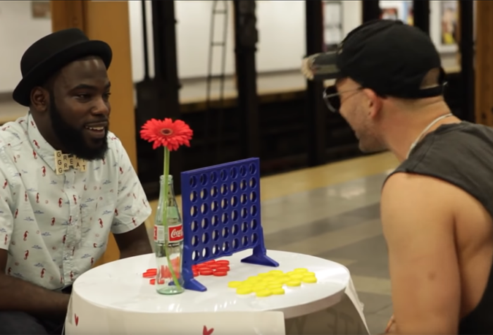 two men sit across small table from each other in subway station, a Connect Four game between them