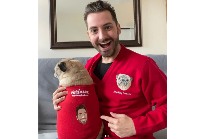 A man wearing a red sweater vest smiles while pointing at his dog, who is also wearing a red sweater vest