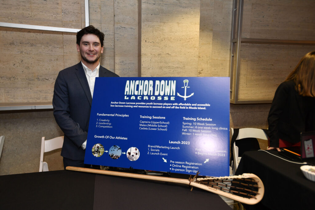 A man stands by a blue board with a lacrosse stick sitting on a table