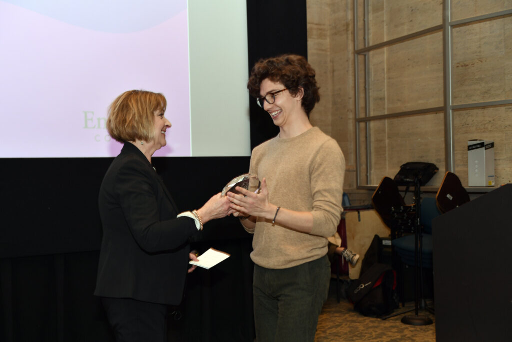 A person smiles gleefully as they accept an award from the program's director