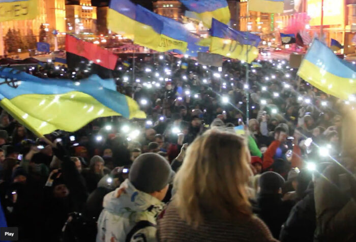people gather in city square at night holding Ukrainian flags and lights
