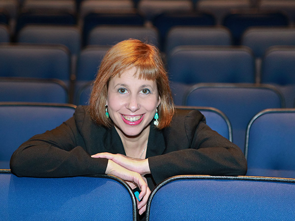Martie Cook in black jacked sitting in empty theater, arms resting on seat in front of her