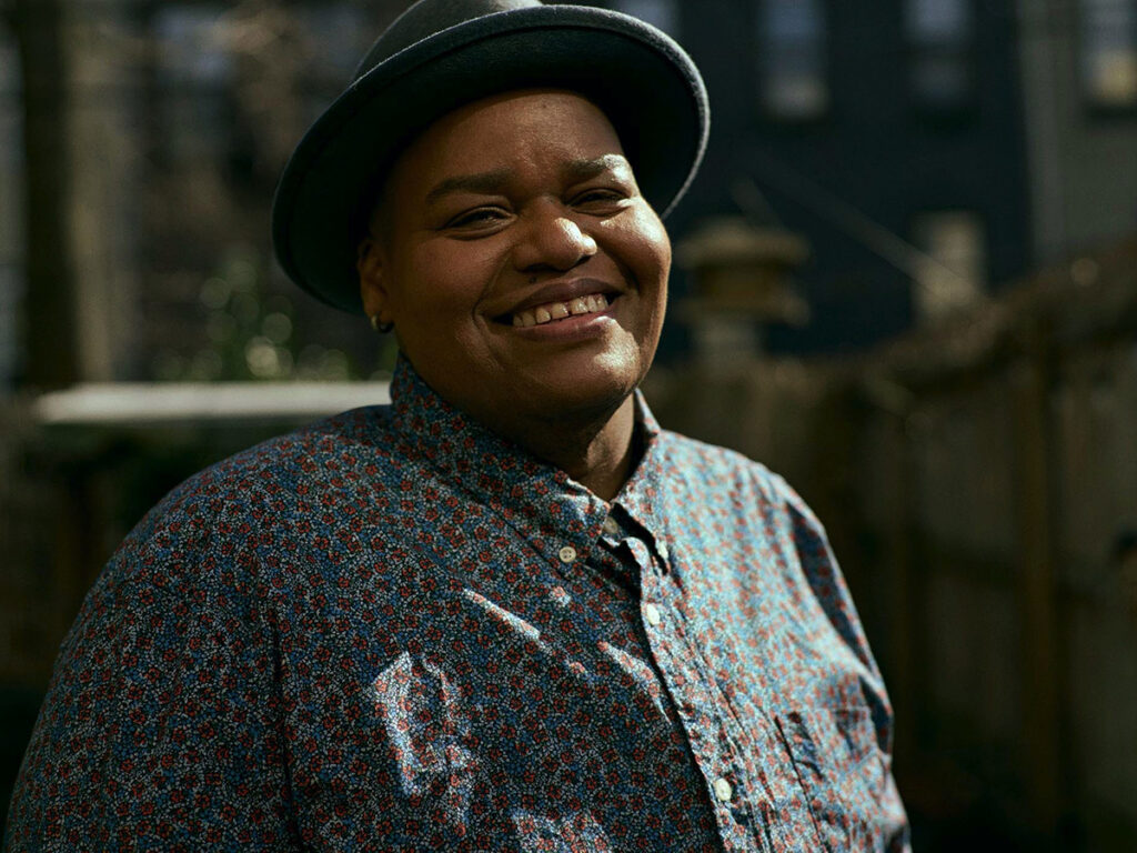 Black woman in brimmed hat, floral shirt smiles with buildings in background