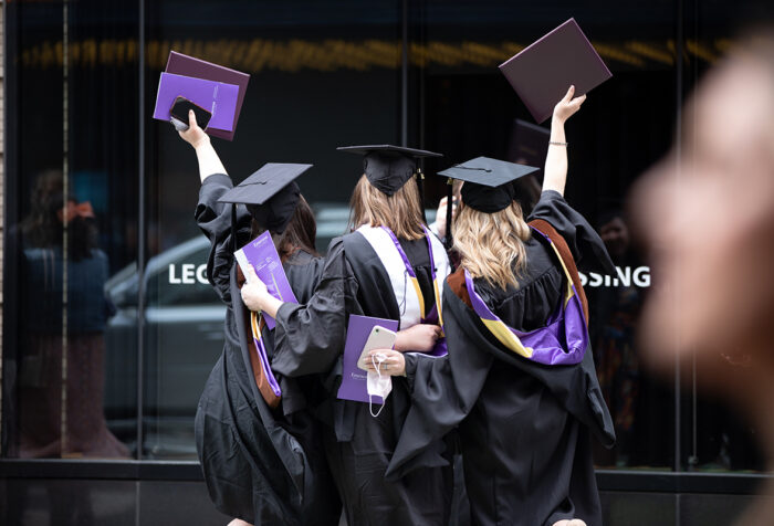 three women in caps and gowns hold up diploma cases with backs to camera