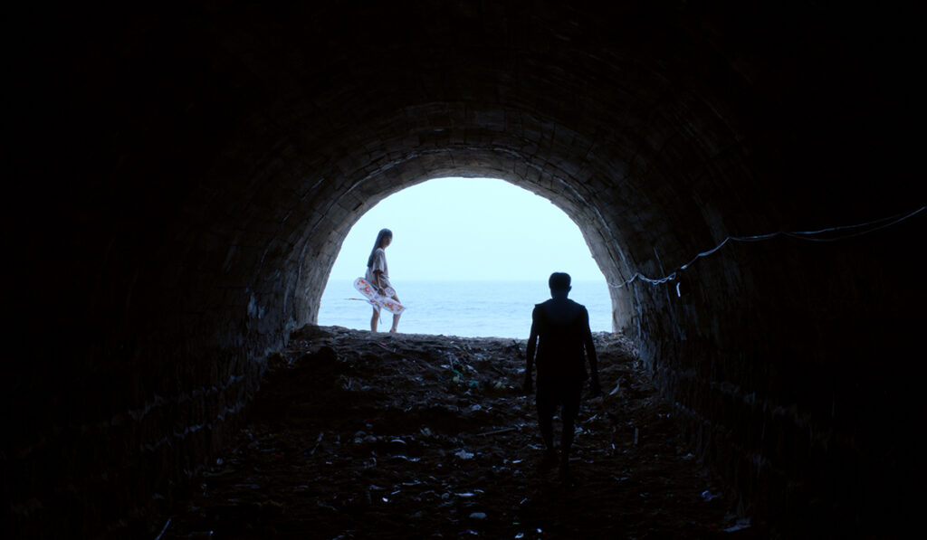 woman stands at end of dark tunnel with sea behind her, man walks toward camera in tunnel