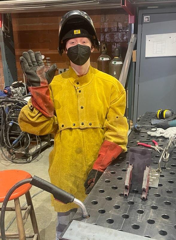 woman in welding mask, yellow apron, work gloves waves at a welding table