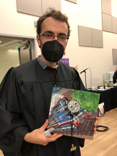A man wearing a graduation gown with a black mask holds a graduation cap with an image of Thomas the Train on it