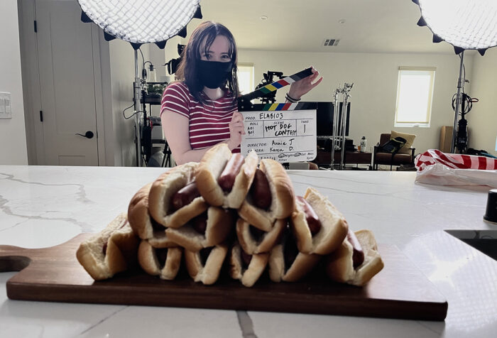 young woman wearing mask holds a clapperboard behind a stack of hot dogs on a table