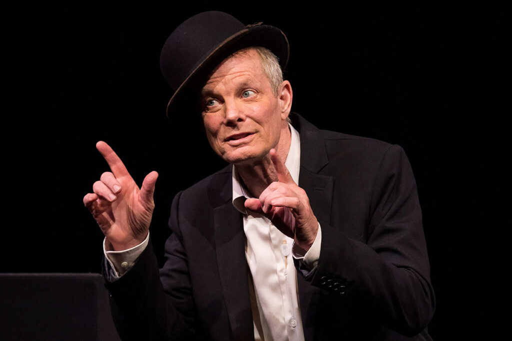 white man in black jacket and bowler hat points gestures with both index fingers