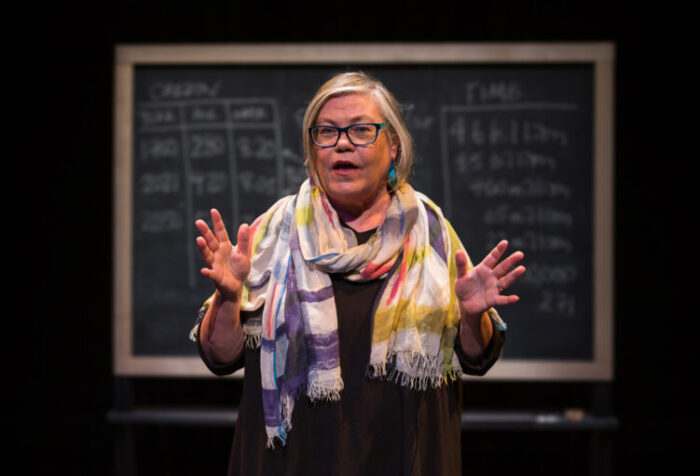 woman in scarf talks on stage in front of blackboard