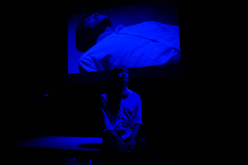 Asian man sits in front of screen showing himself lying prone, all bathed in blue light
