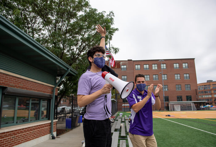 young man in glasses, mask, and purple T-shirt speaks into megaphone with arm raised as another man looks on and claps at a ball field