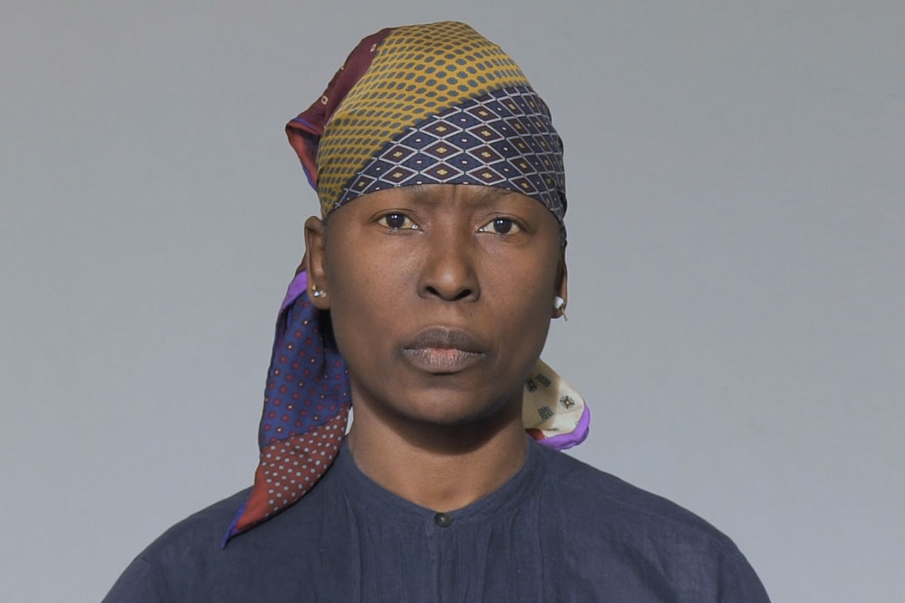 black woman in colorful head scarf looks directly at camera in front of grey background