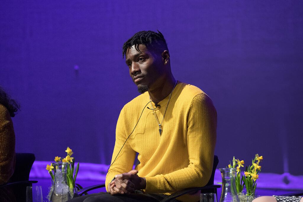 Man in yellow shirt sits on stage against purple background with daffodils around him. 