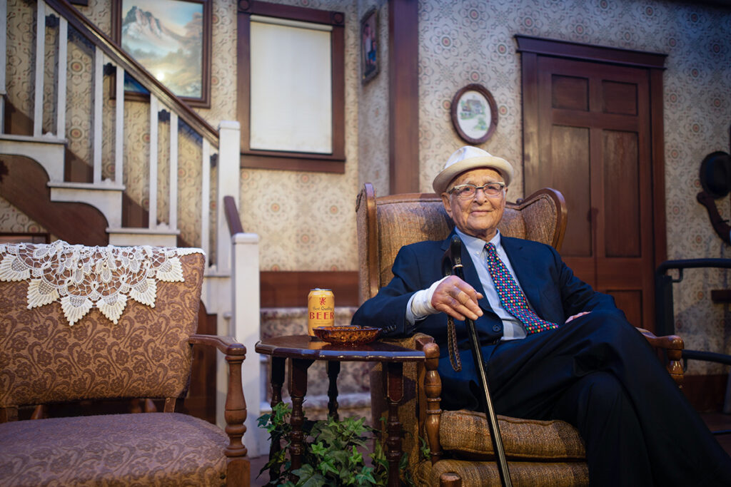 Norman Lear in suit sits in replica set from All in the Family