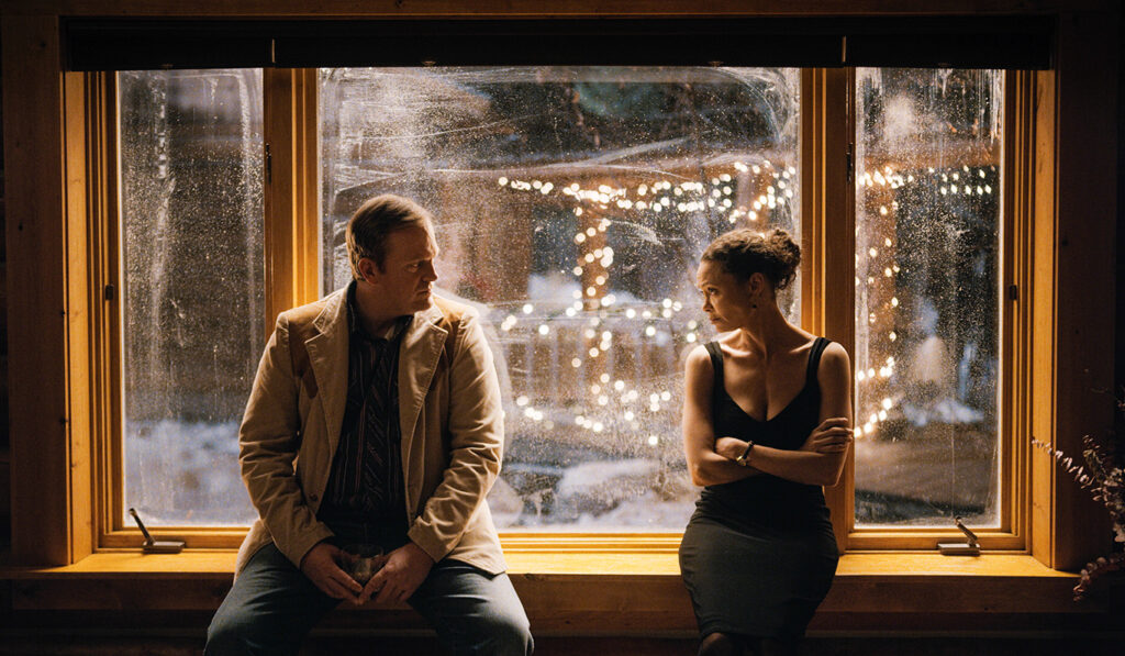 Man and woman sit in window sill looking at each other. White lights outside window
