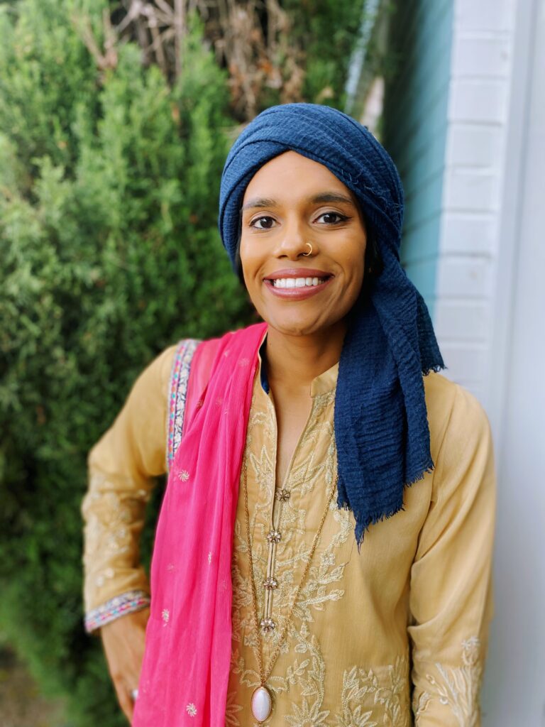 Amber Hai in tan top, blue headscarf leans against wall with a shrub behind her