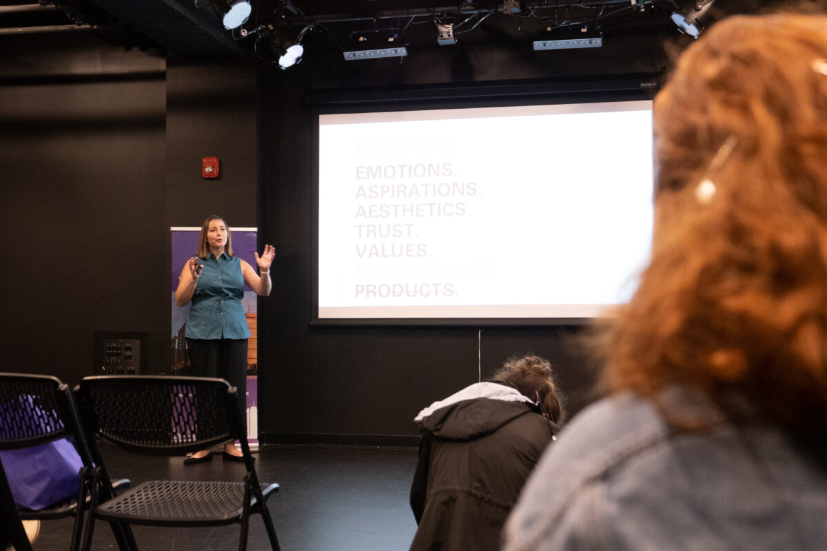 Brenna McCormick stands to the left of the screen, gesturing to the crowd. On the screen are the words "emotions, aspirations, aesthetics, trust, values and products."
