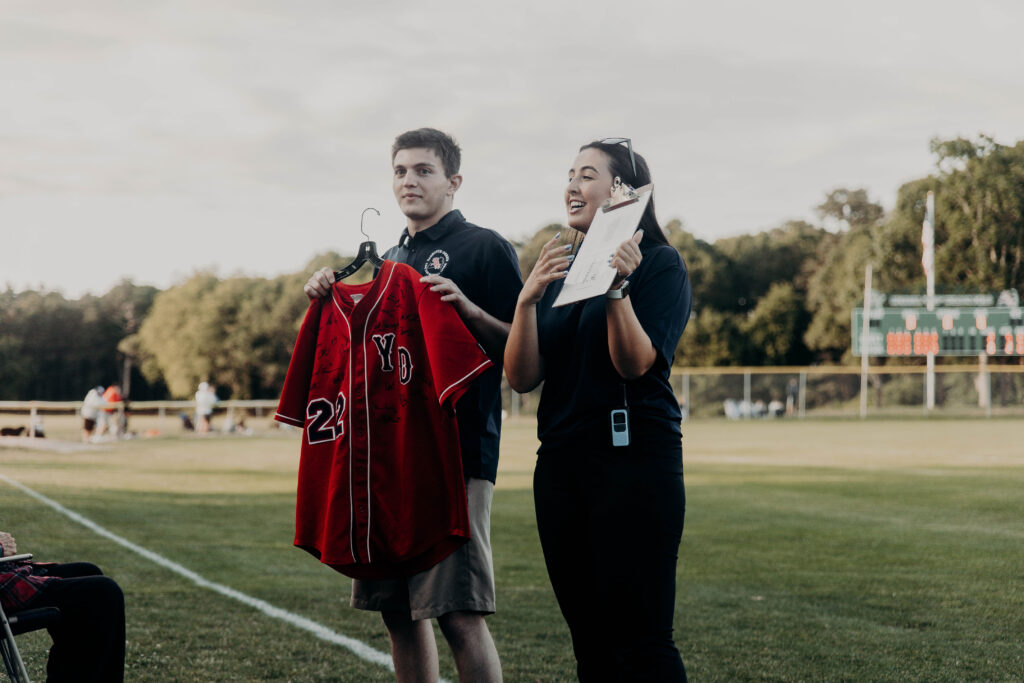 As a Gameday Operations intern brining his game to the fan experience, Maximilian Mandel '24 stands on the edge of a green baseball field facing the stands as he holds up a red jersey with the number 22 signed by all the players. [Courtesy Photo] 
