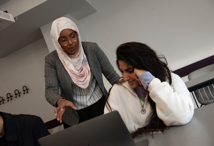 Woman in hijab talks to and looks over the shoulder of a woman seated in front of open laptop