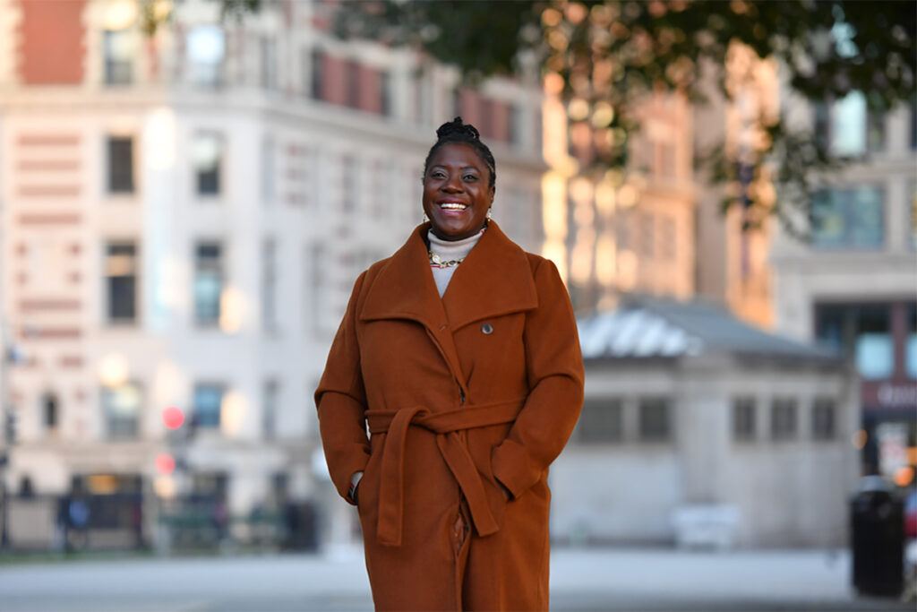 Shaya in rust colored coat, smiling and standing in Boston Common, Boylston T station and buildings behind
