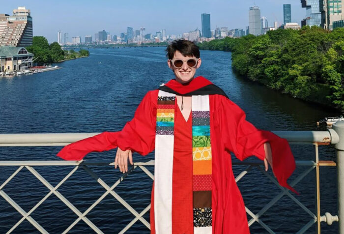woman in red academic robes and rainbow hood leans against railing of bridge, Charles River and Boston skyline in distance