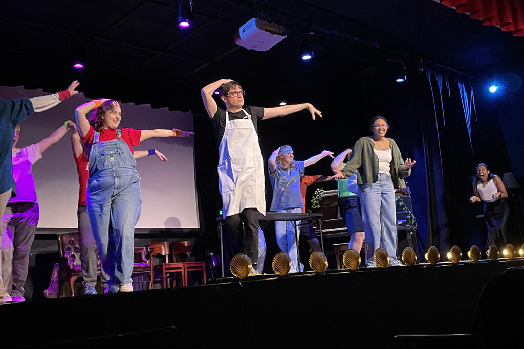 man wearing white apron dances on stage surrounded by others doing same move