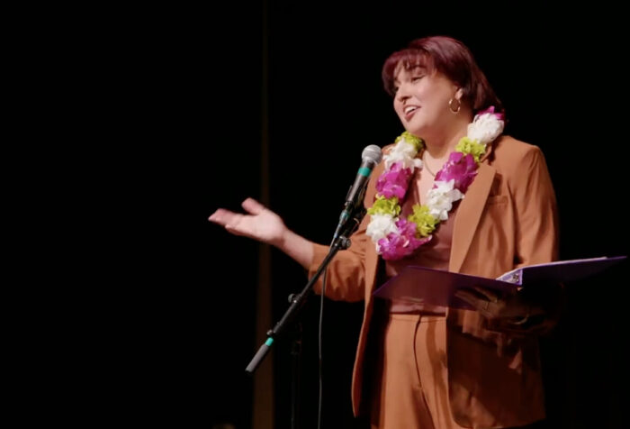 woman in brown suit, lei, speaks on darkened stage
