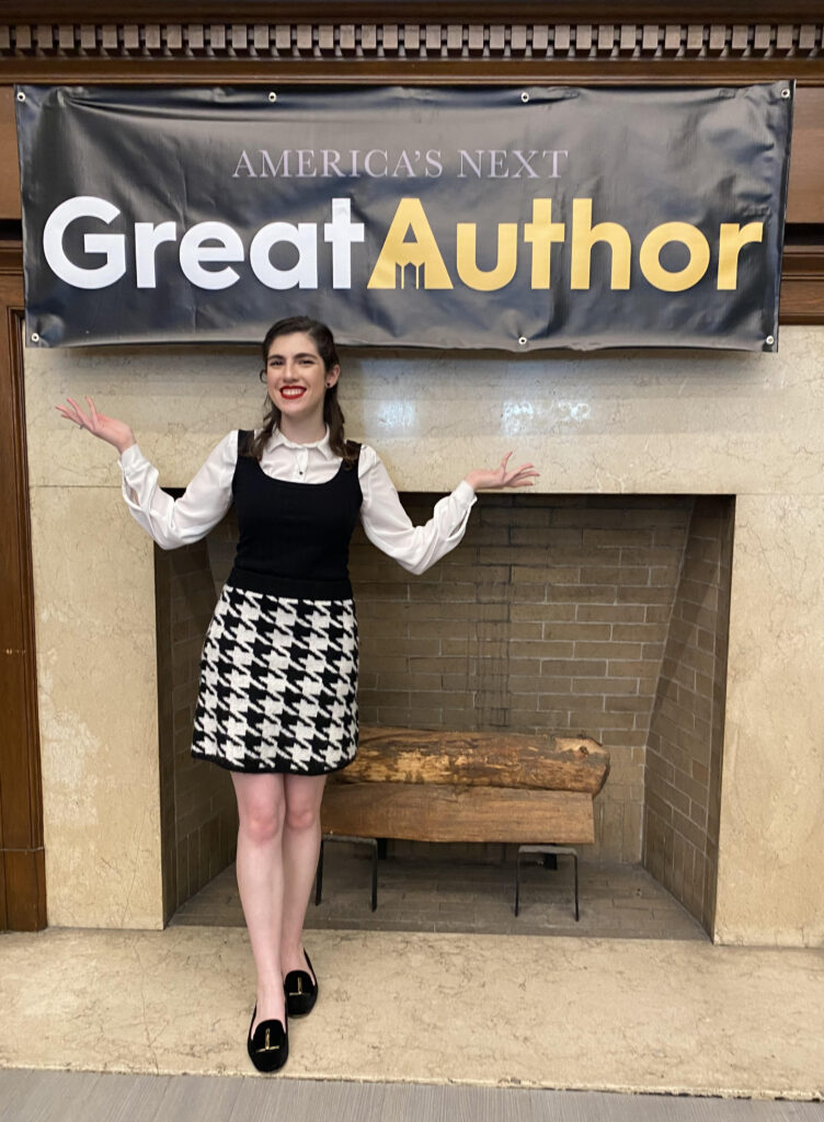 woman in white shirt, black and white houndstooth jumper stands in front of fireplace and banner reading "America's Next Great Author"