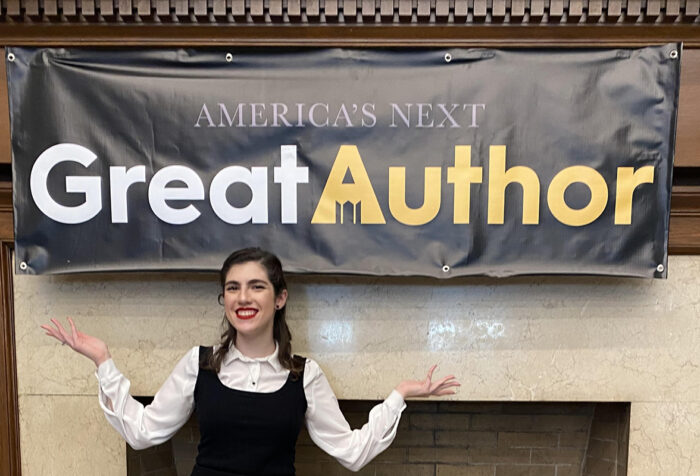 woman in white shirt, black jumper stands in front of fireplace and banner reading 'America's Next Great Author'