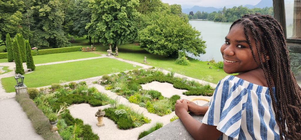young woman on balcony looks over shoulder, gardens, lake, and Austrian mountains in background