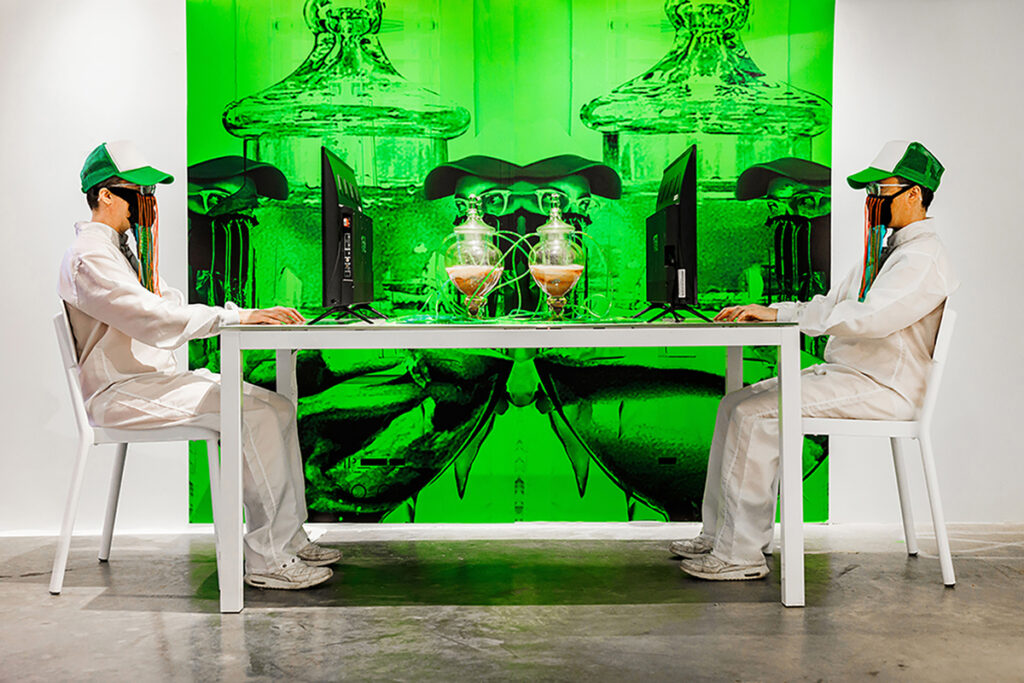 two men in white jumpsuits, trucker hats, wearing masks, sit at computer screens across table with jars filled with unidentified substance in center. on wall behind is a green tinted photograph of the jars