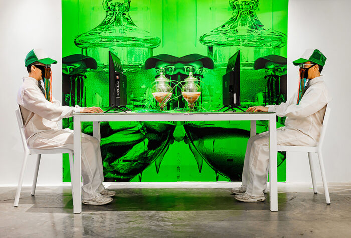two men in white jumpsuits, trucker hats, wearing masks, sit at computer screens across table with jars filled with unidentified substance in center. on wall behind is a green tinted photograph of the jars