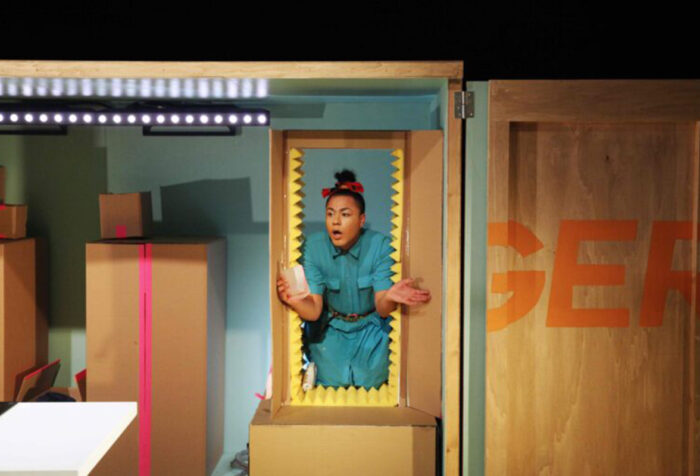 A trans woman in a blue jumpsuit, red head scarf, pops out from cardboard box, on set full of cardboard boxes