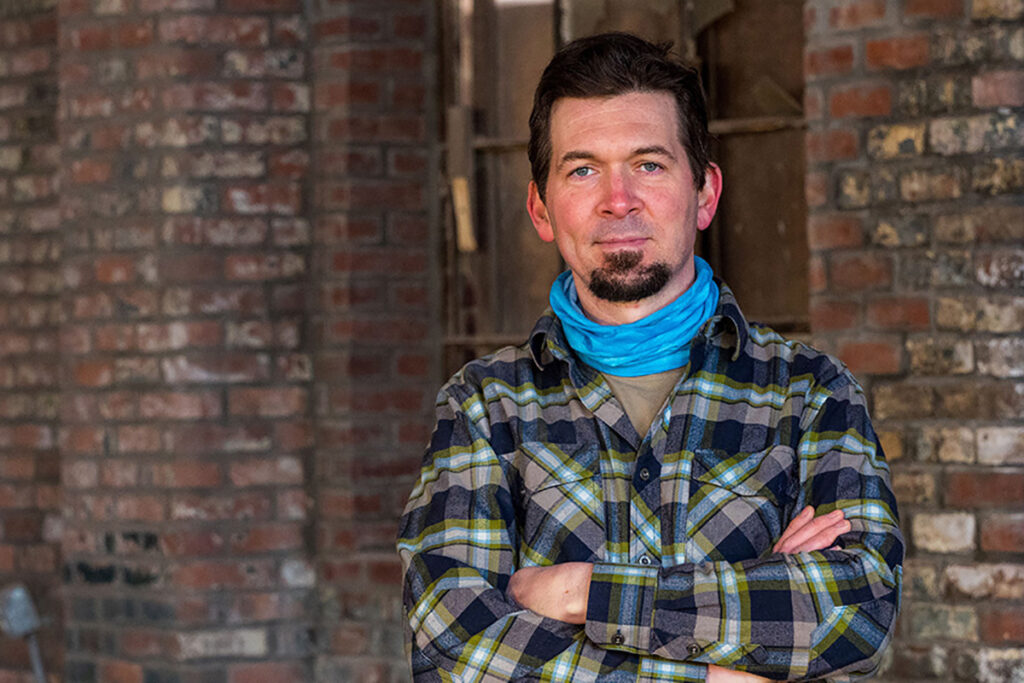 man in plaid shirt stands with arms crossed in front of brick building