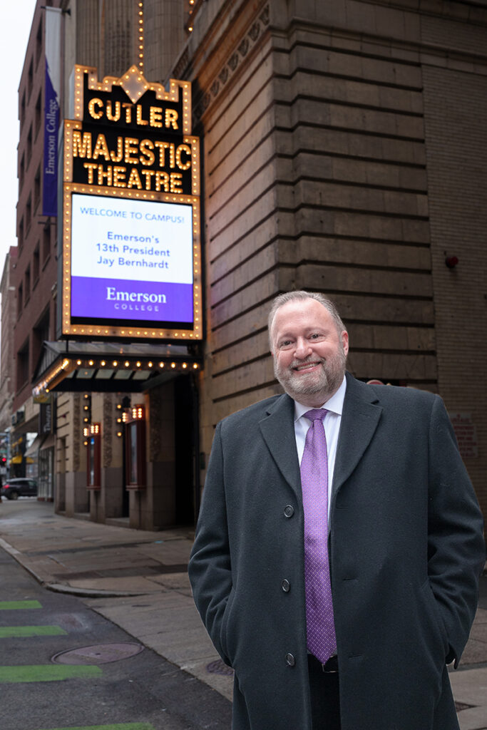 Man in suit coat and purple tie stands in front of theater marquee that reads "Welcome to Campus! Emerson's 13th President Jay Bernhardt" 