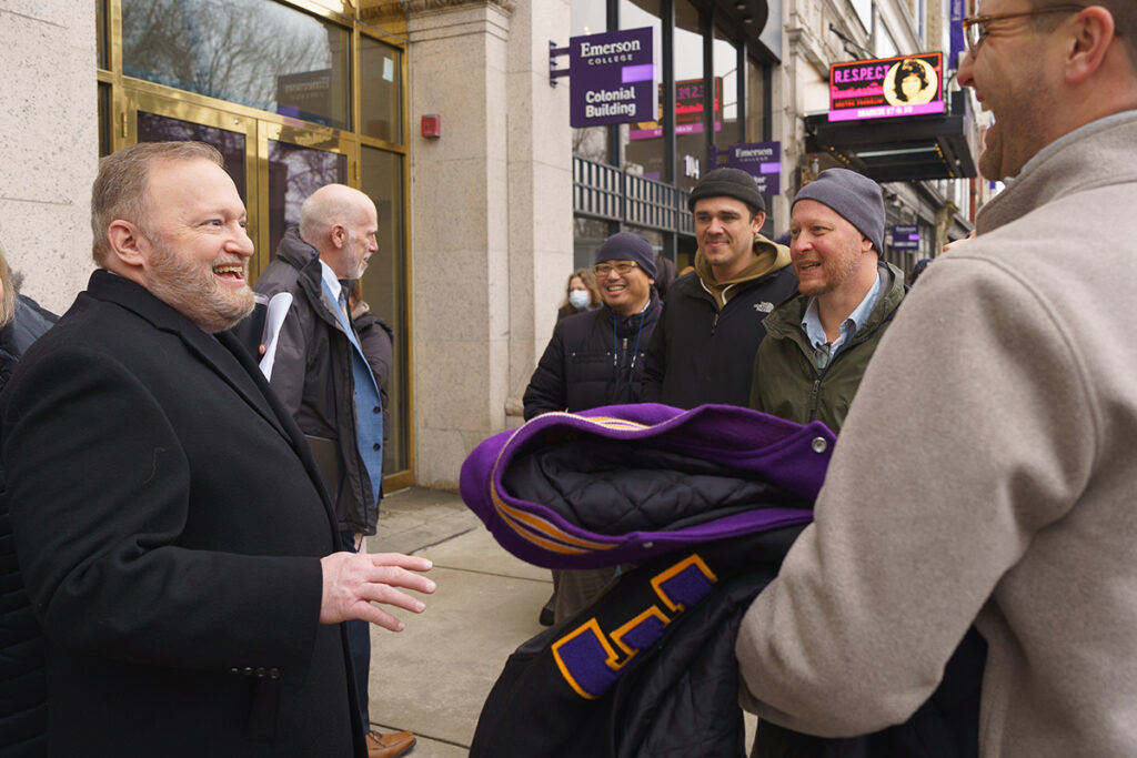 jay bernhardt, in dark coat, smiles and chats with Emerson employees in front of the Colonial Building. Employee closest to camera holds a purple and gold varsity jacket