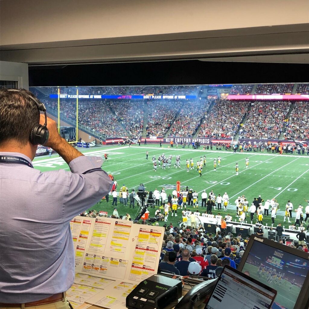 Bob Socci uses binoculars to look at a football game from his broadcast box