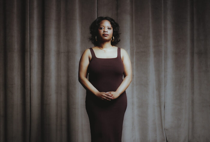 Black woman in brown dress stands with hands clasped against beige curtains